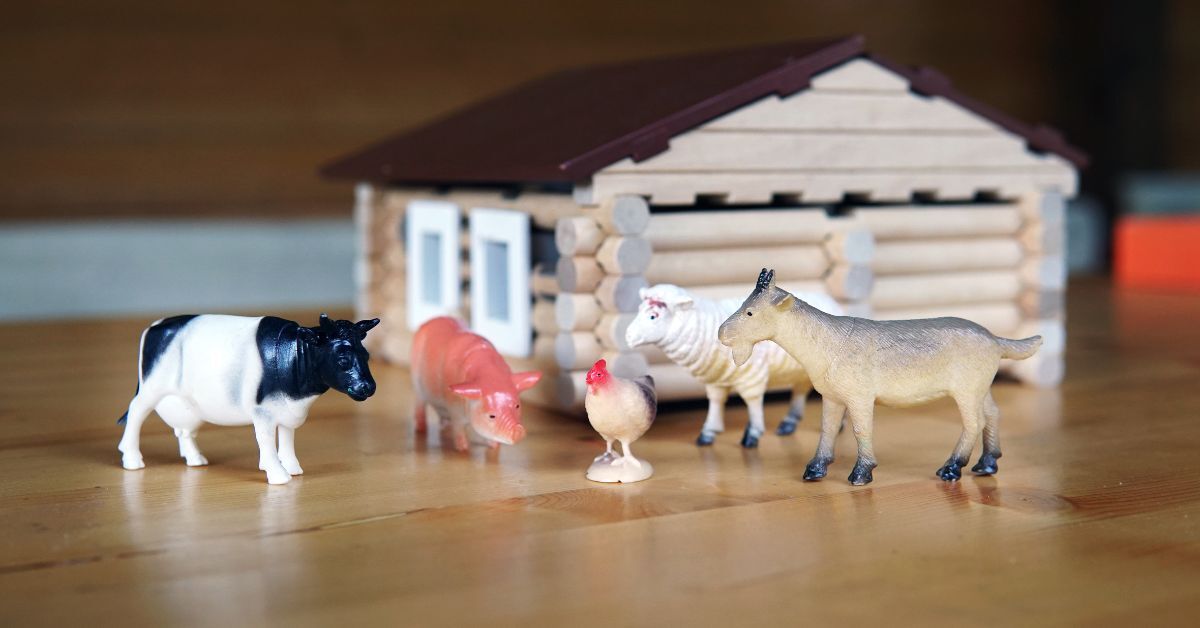 A plastic cow, pig, chicken, sheep, and goat set up in front of a small toy log cabin on a wood floor.