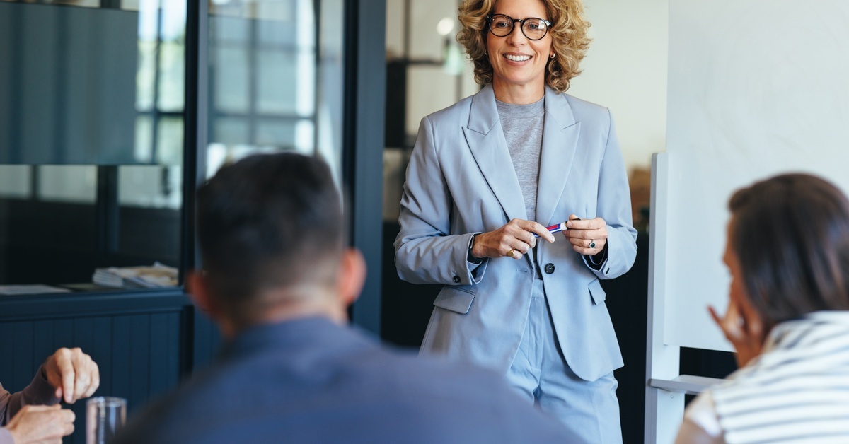 A woman in a gray suit and glasses smiles and holds a pen while she looks at her two seated coworkers.