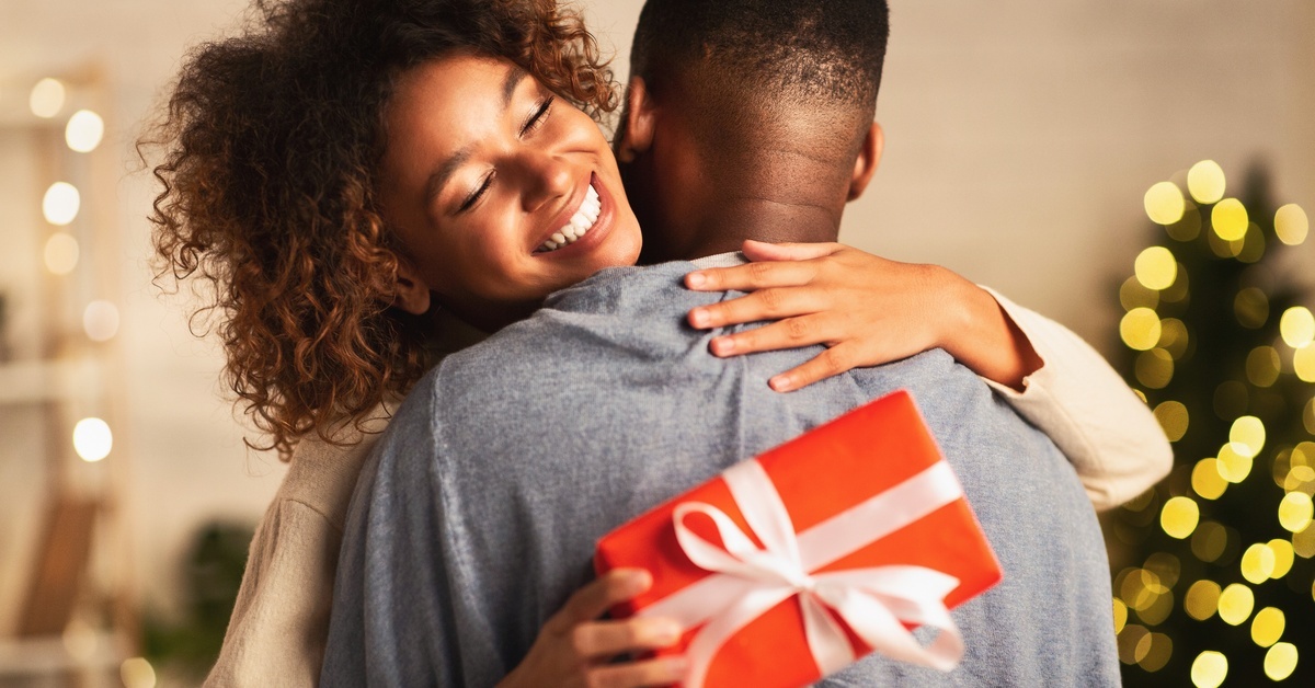 The Art of Giving: Why Small Unique Gifts Mean So Much A woman holding a small, red gift box with a white ribbon around it. She smiles as she hugs a man in a gray shirt.