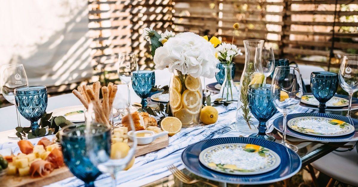 An outdoor table set with blue and white plates and glasses, with flowers and lemons spread out as decor.
