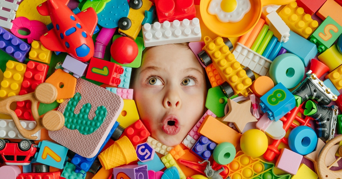A young child's surprised face emerges from a pile of toys with building blocks, miniature cars, animals, and other objects. A young child's surprised face emerges from a pile of toys with building blocks, miniature cars, animals, and other objects.