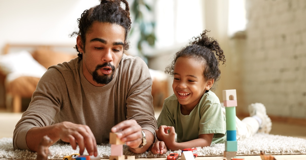 A dad and his child lay on the floor on a carpet, playing with blocks, miniature cars, and animals. A dad and his child lay on the floor on a carpet, playing with blocks, miniature cars, and animals.