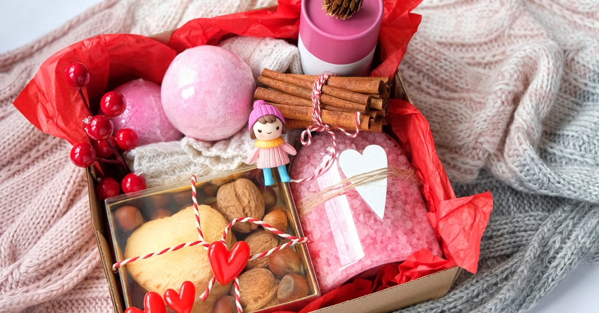 A gift box full of small self-care items including bath bombs, salts, a box of snacks, and a small doll in the center. A gift box full of small self-care items including bath bombs, salts, a box of snacks, and a small doll in the center.