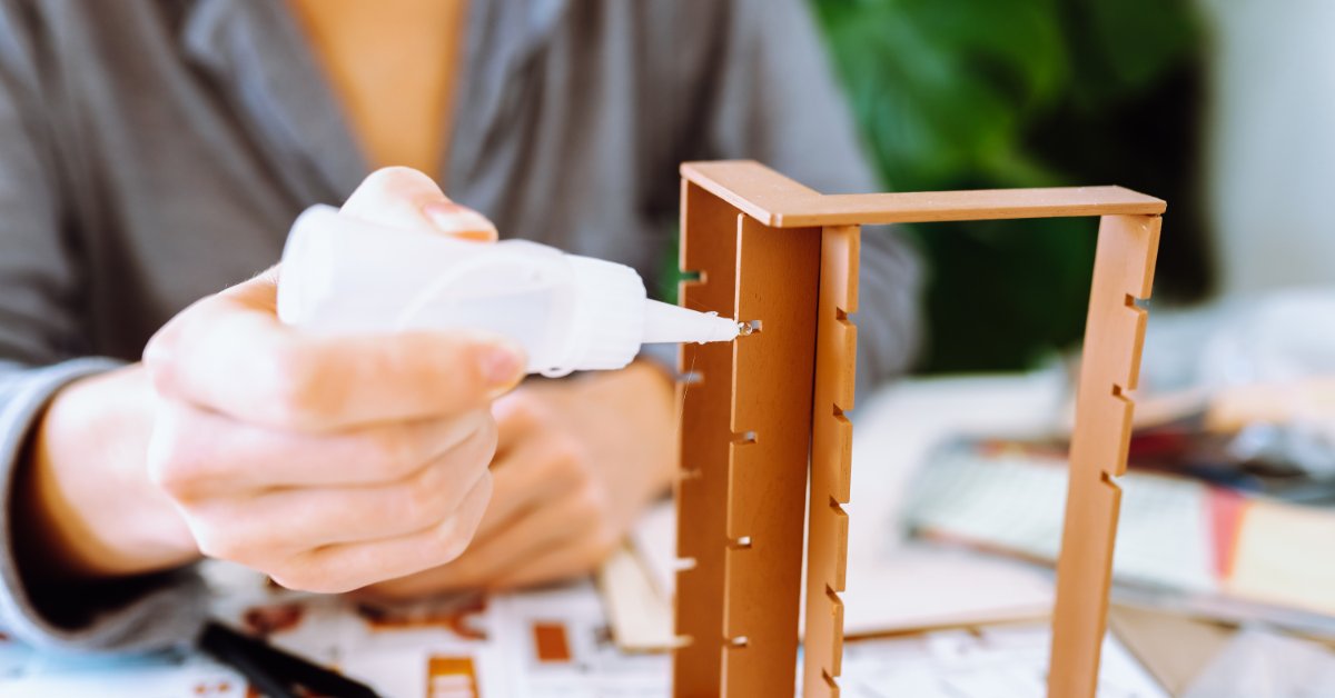 Hands applying glue while assembling a wooden house model on a table with small craft pieces and plants nearby.